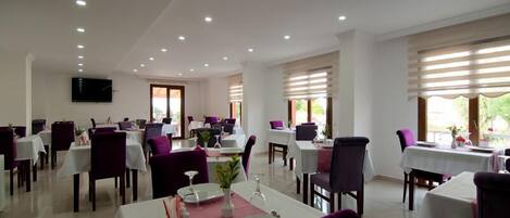 A dining area with tables set for two, white tablecloths, and purple chairs.