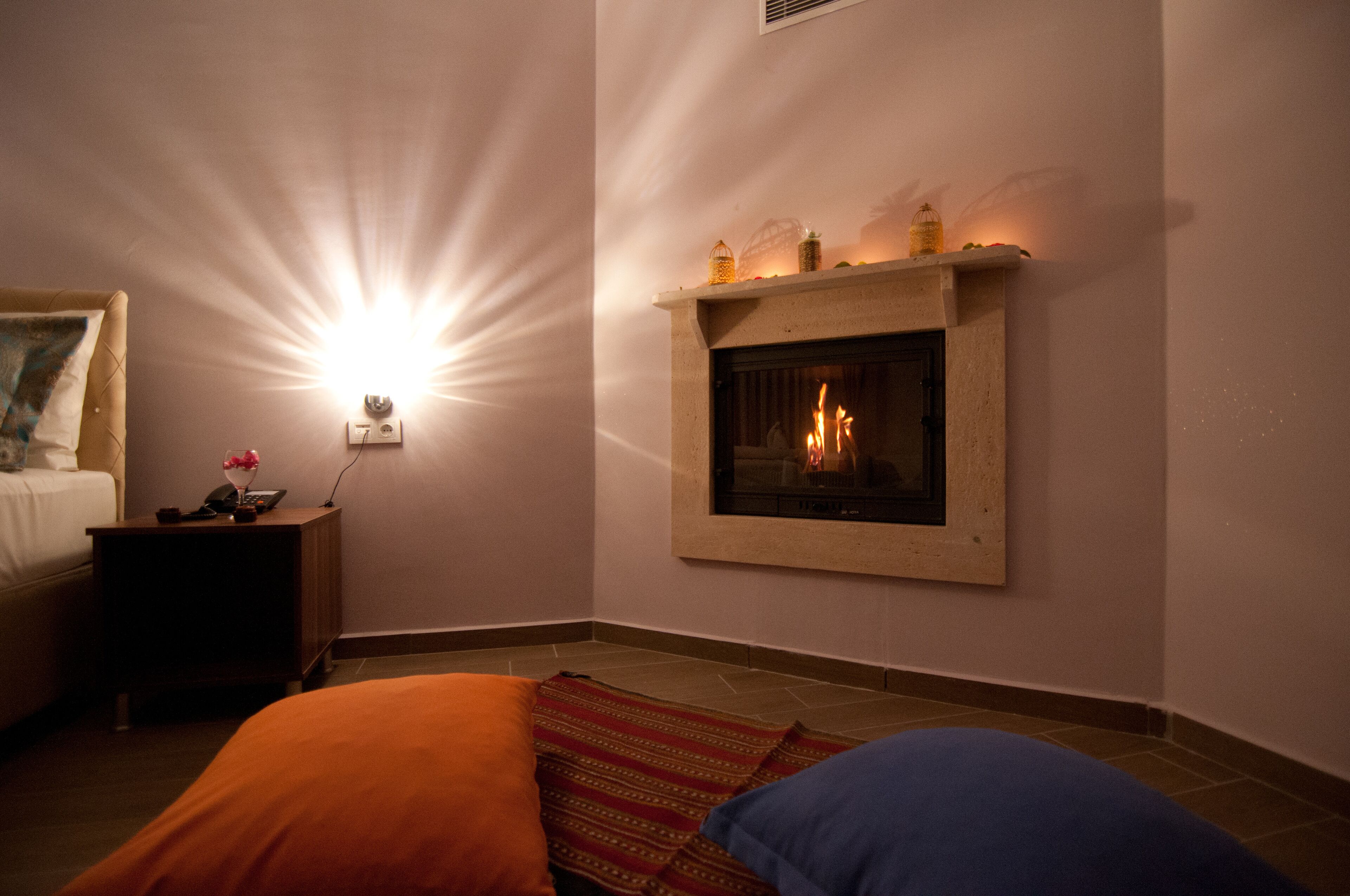 Una camera d'albergo con camino, un letto con cuscino arancione e blu, un comodino con telefono e un vaso di fiori, e una lampada a parete.