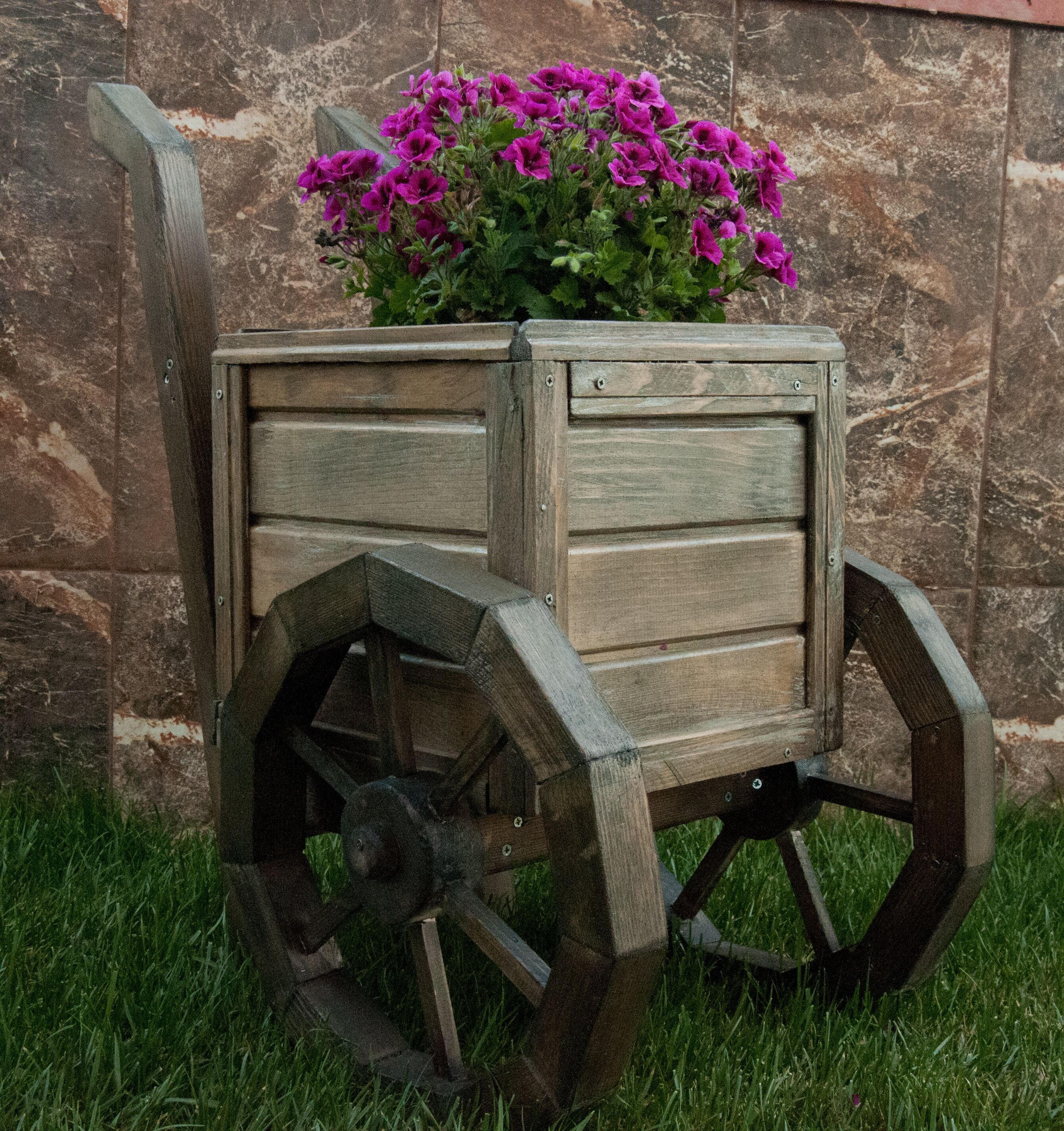A wooden cart with wheels and a flower pot on top, placed on grass against a stone wall.