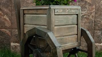 A wooden cart with wheels and a flower pot on top, placed on grass against a stone wall.