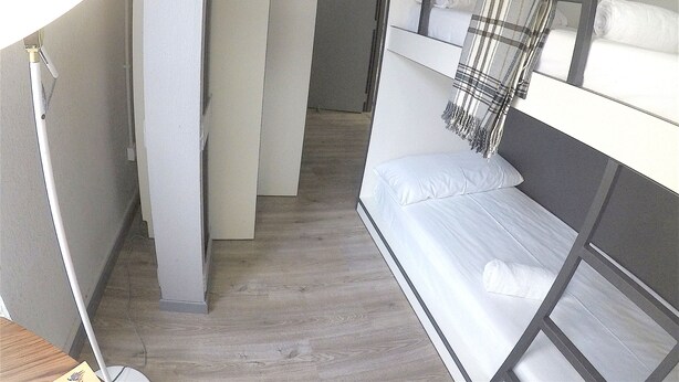 A narrow, dimly lit corridor with bunk beds on one side and a white lamp on the other. - Quartier Bilbao Hostel (Bilbao)