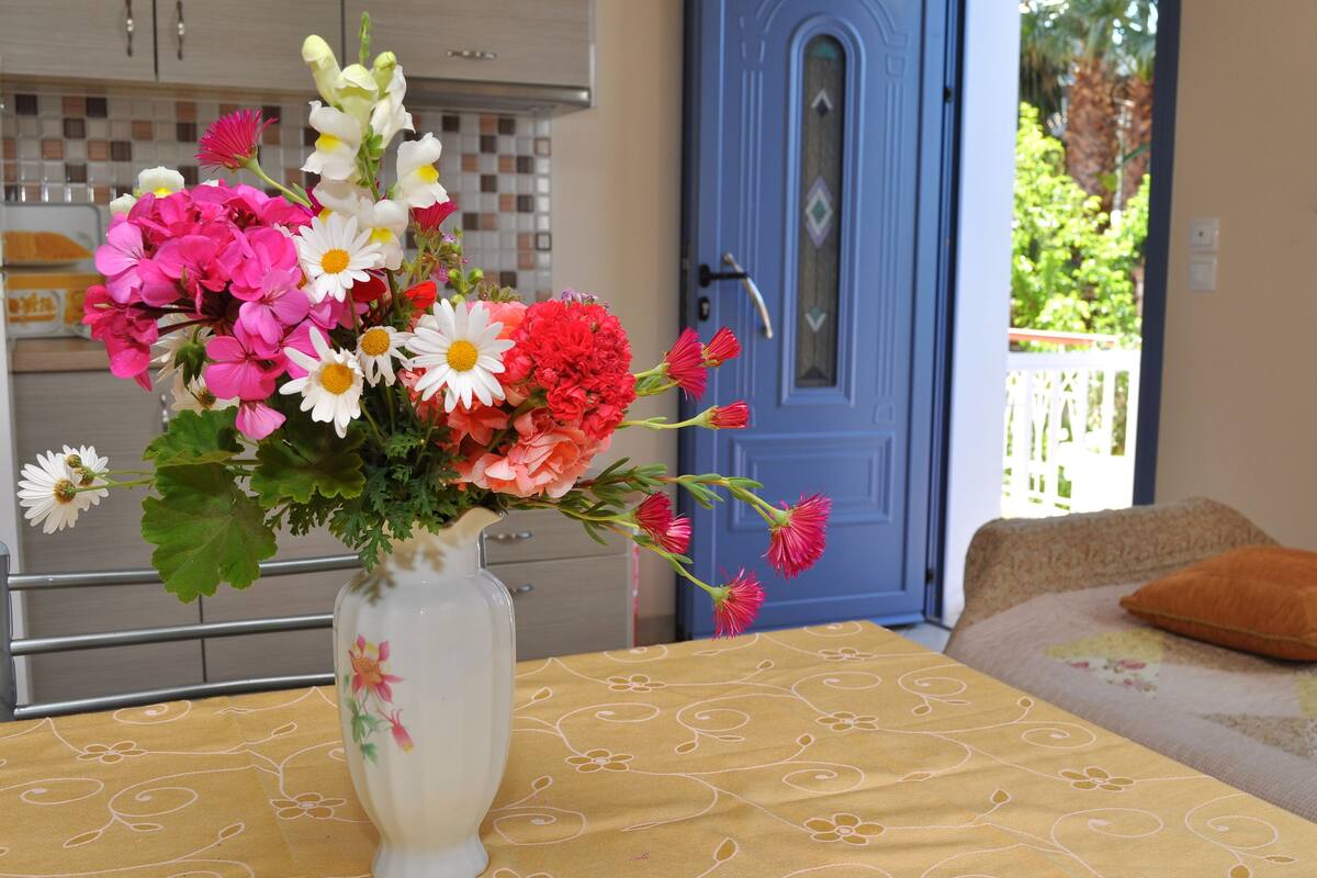 Ein Esszimmer mit gedecktem Tisch, einer Blumenvase und einer blauen Tür, mit Blick auf eine grüne Landschaft.