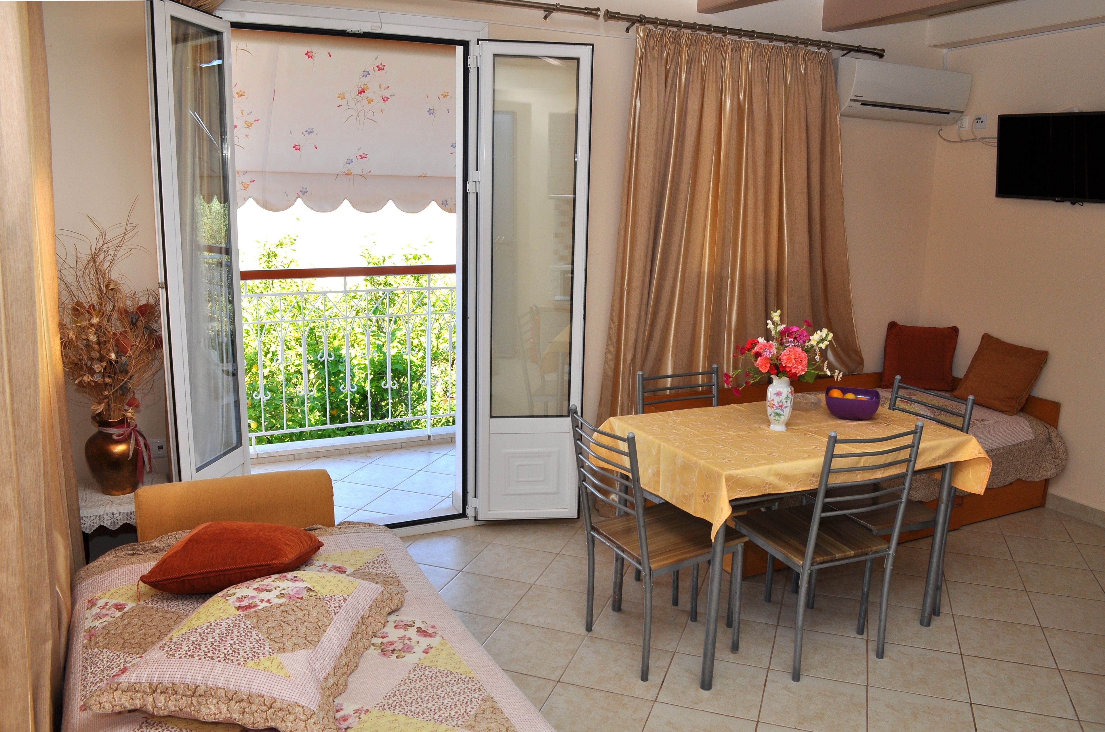A dining area with a table set for two, a sofa with cushions, and a view of a green garden through an open door.