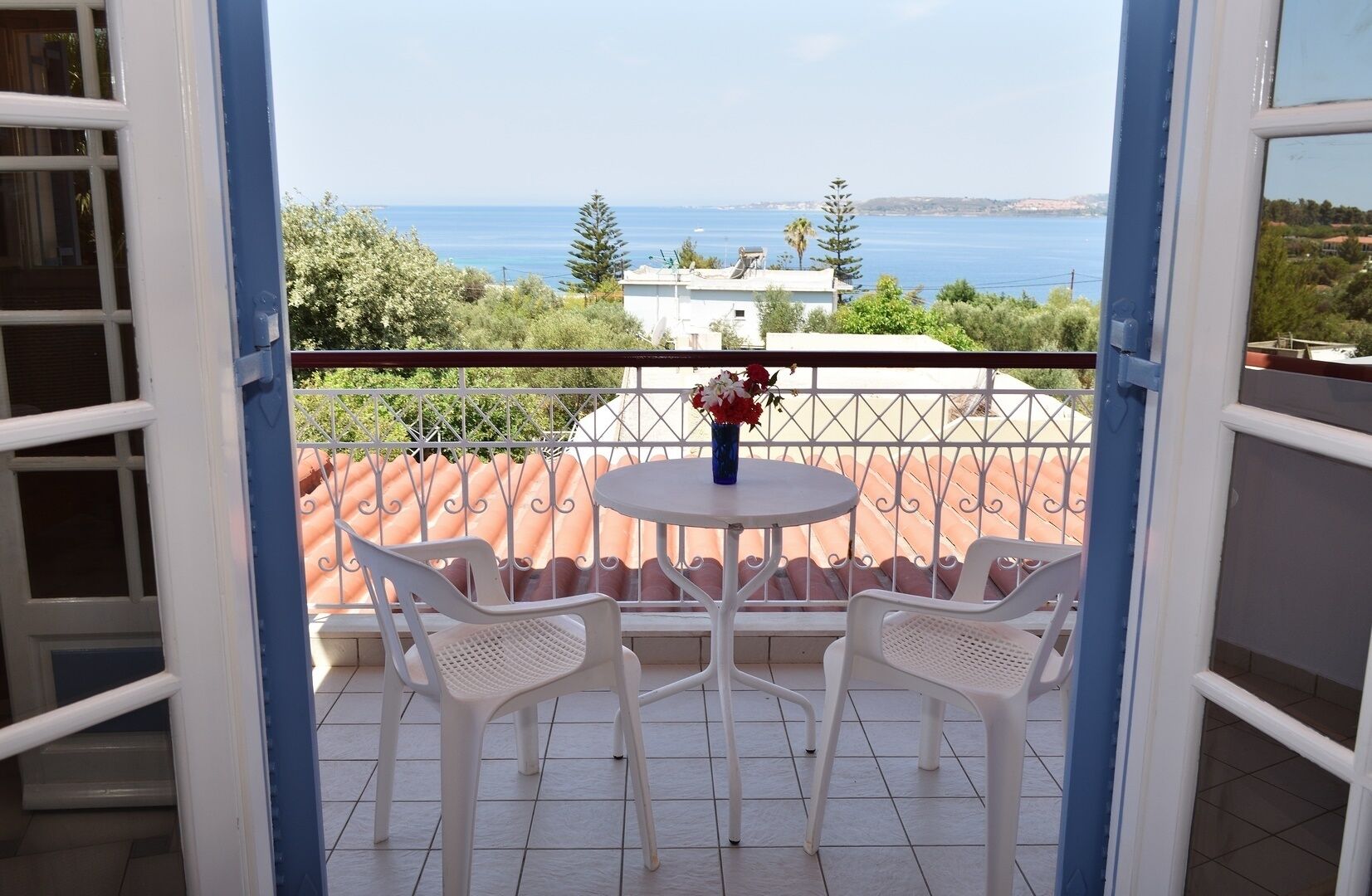 A balcony with a table and two chairs, overlooking a coastal view with buildings and trees.