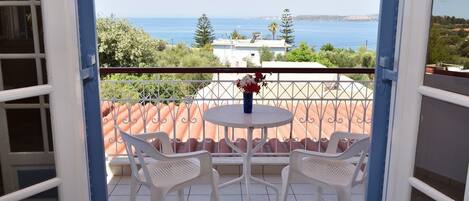 A balcony with a table and two chairs, overlooking a coastal view with buildings and trees.