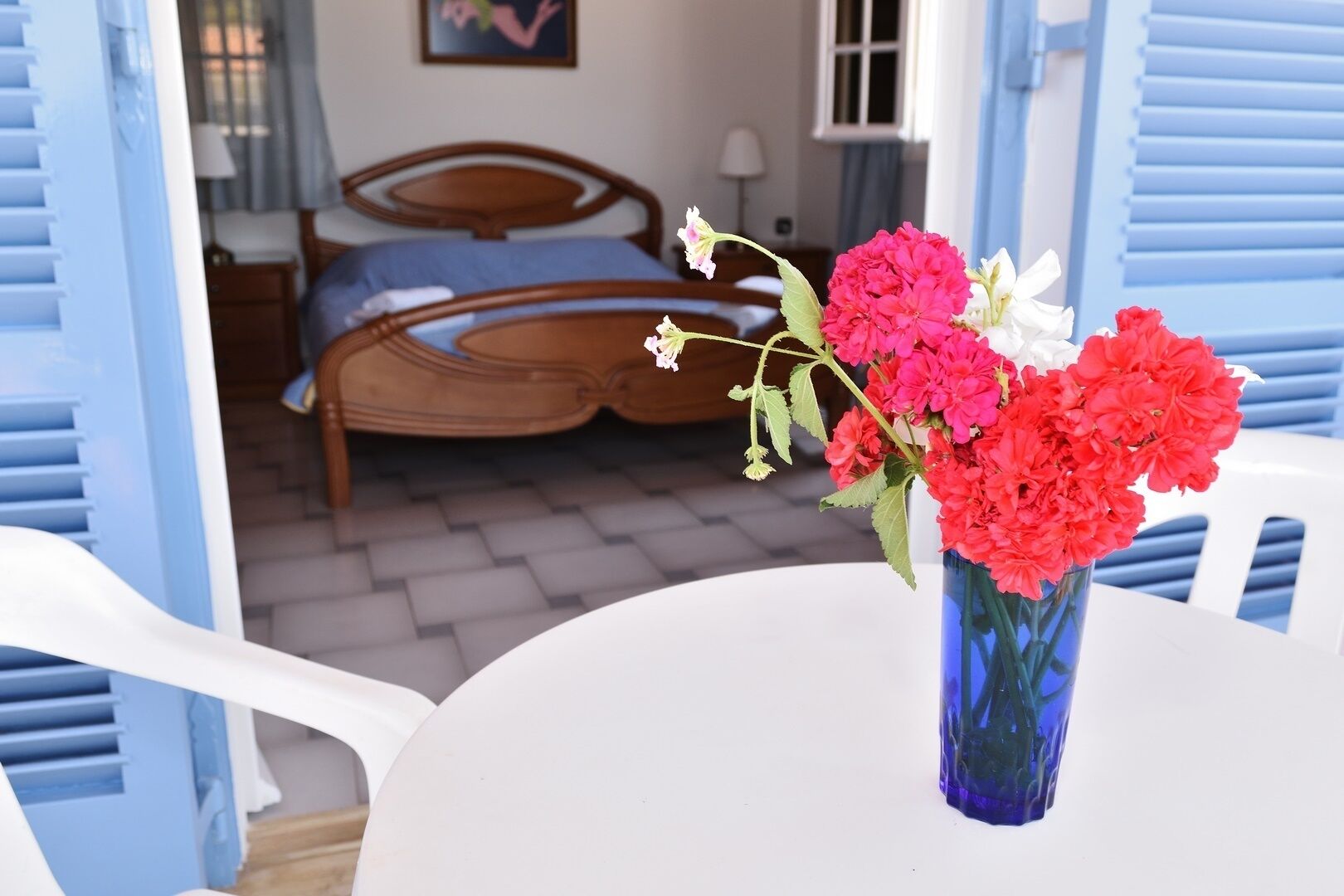 A room with a bed, a table with a vase of flowers, and blue shutters.