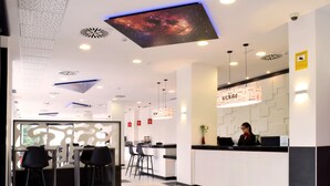 A modern hotel reception area with a digital ceiling display, a reception desk, bar seating, and a staircase in the background. - Hotel New Bilbao Airport (Derio)
