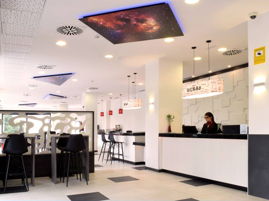 Un área de recepción moderna de hotel con una pantalla digital en el techo, un mostrador de recepción, asientos de bar y una escalera al fondo.