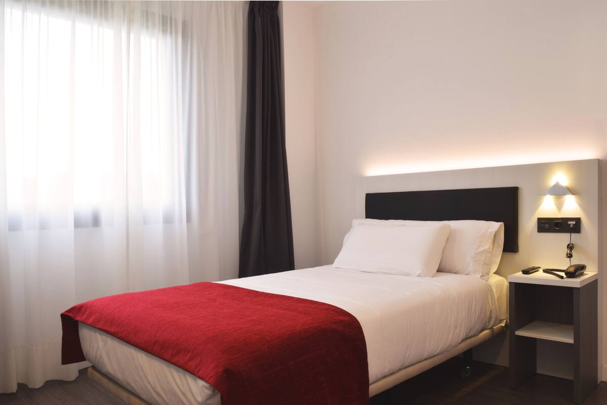 Una habitación de hotel con cama, mesita de noche, lámpara y ventana con cortinas.