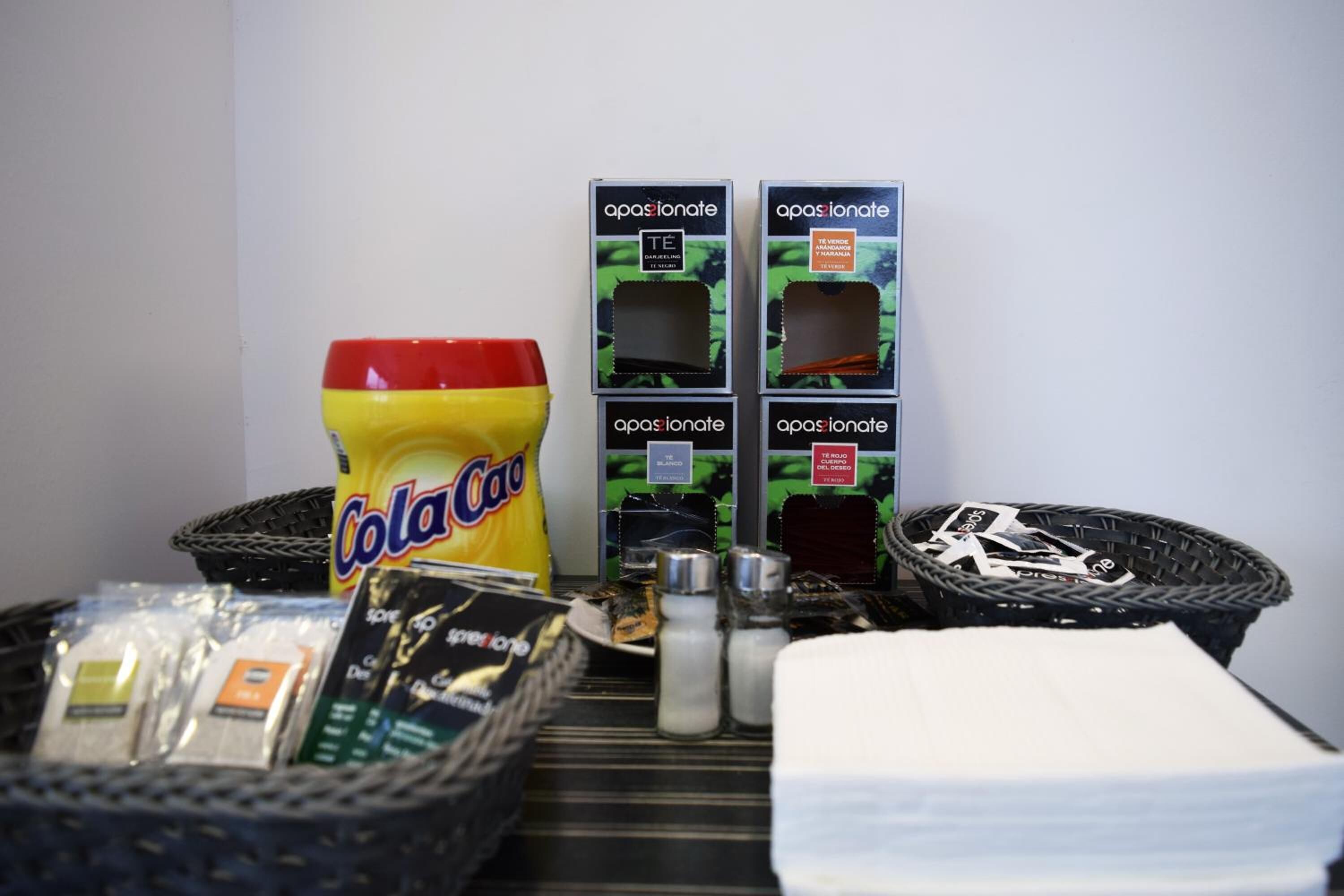 Una mesa con varios objetos, incluyendo una botella de Coca-Cola, cajas de productos 'apasionate', una cesta con sobres y una pila de platos blancos.