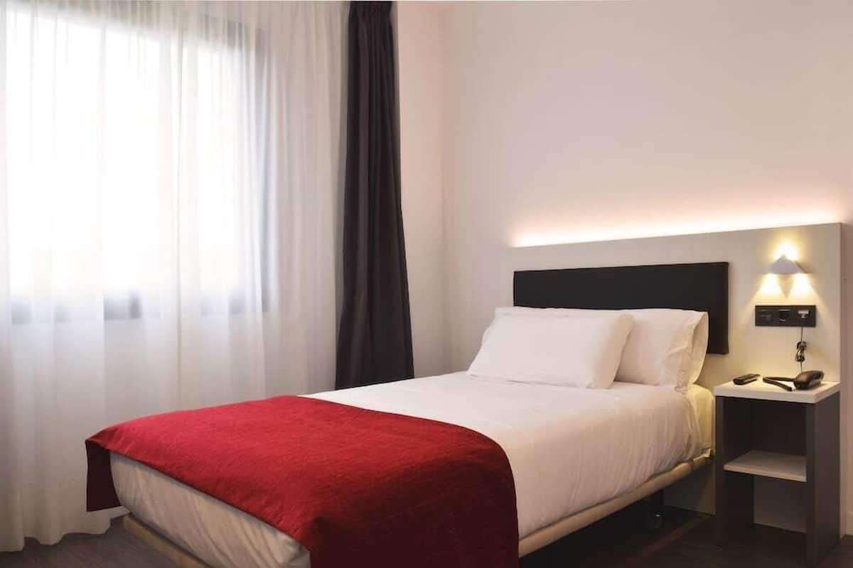 Una habitación de hotel con una cama, una mesita de noche con lámpara y una ventana con cortinas.