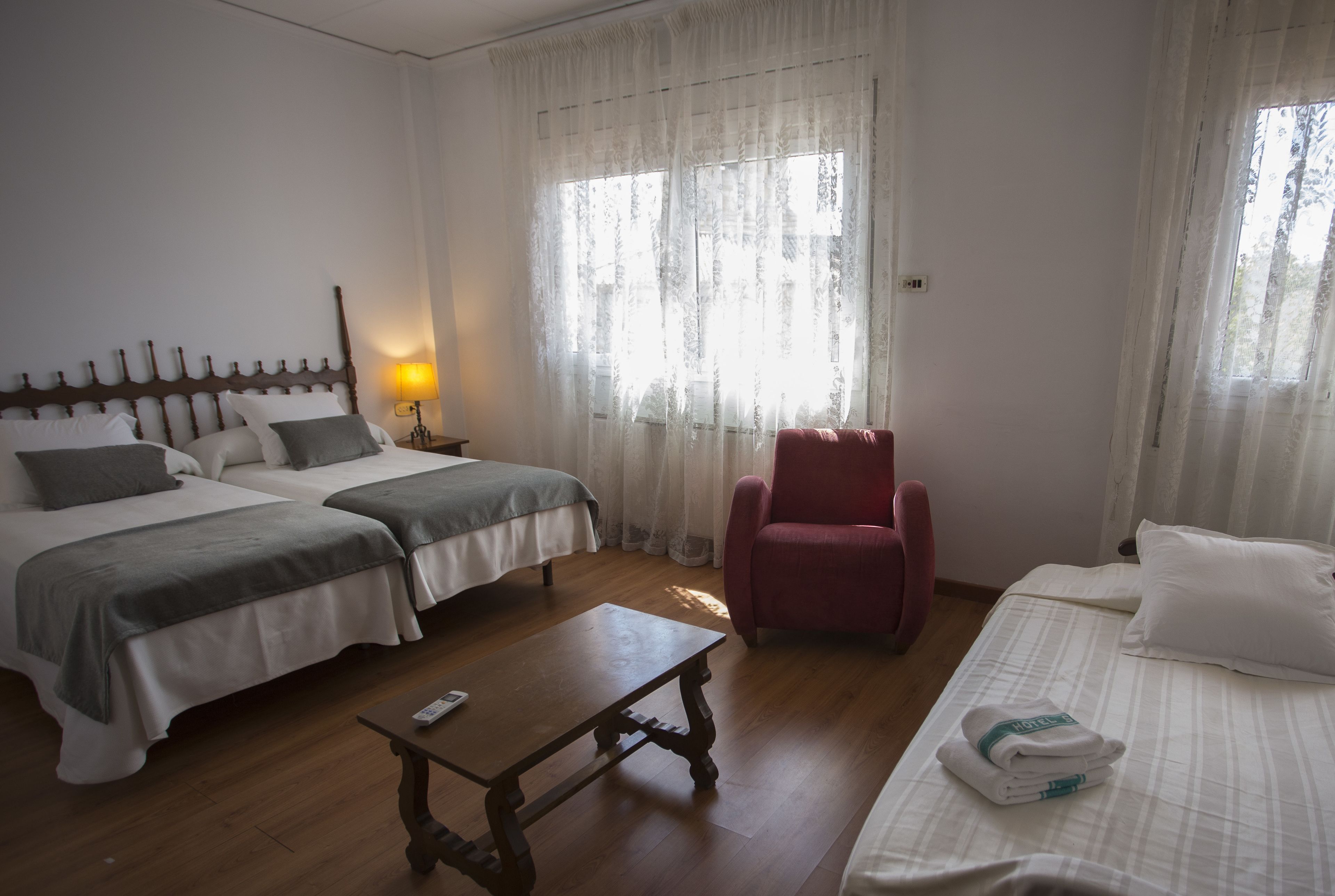 Habitación de hotel con dos camas, una mesa de madera, una silla roja y una ventana con cortinas traslúcidas.