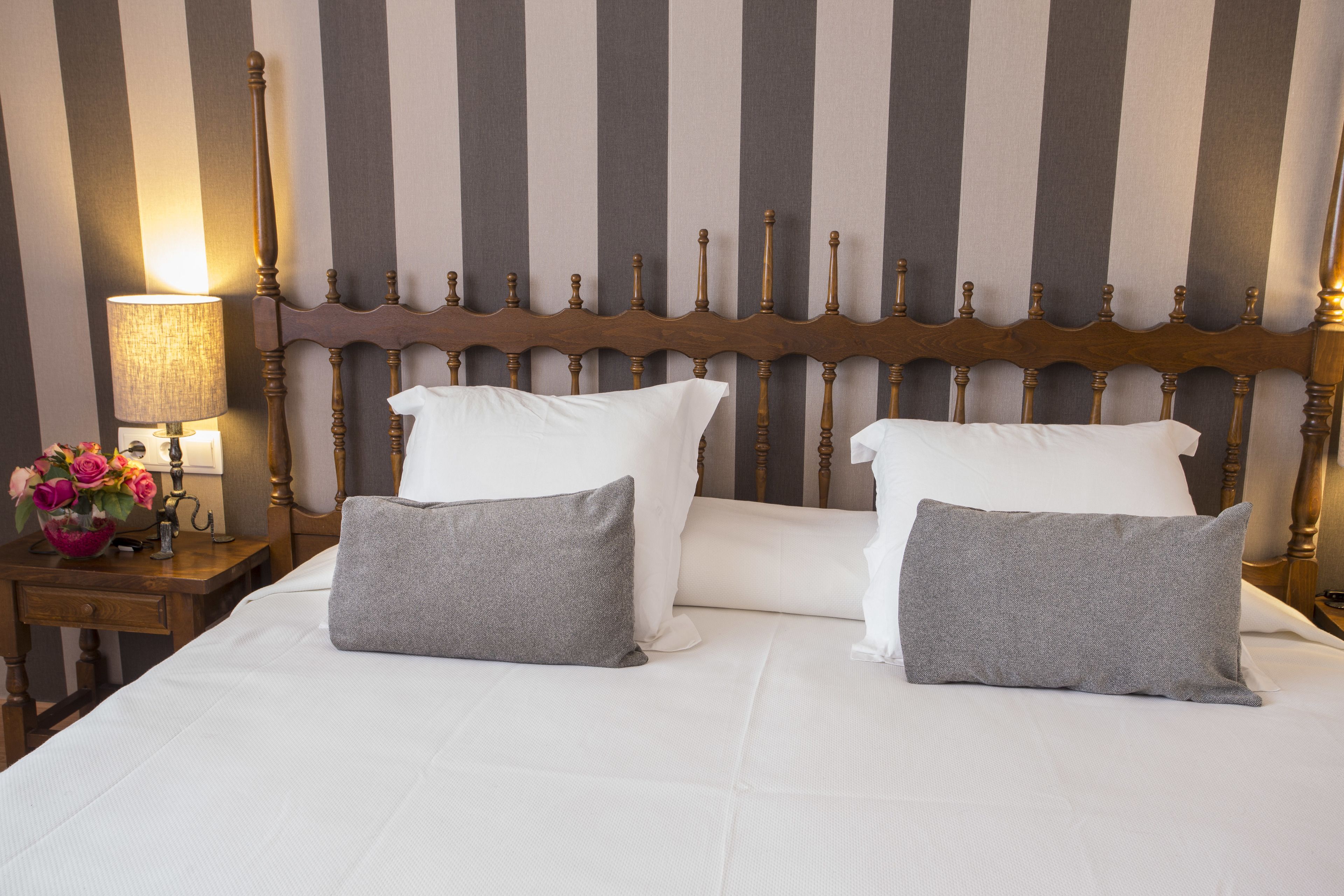 Una cama bien hecha con almohadas blancas y grises, un cabecero de madera y una mesita de noche con una lámpara y flores.