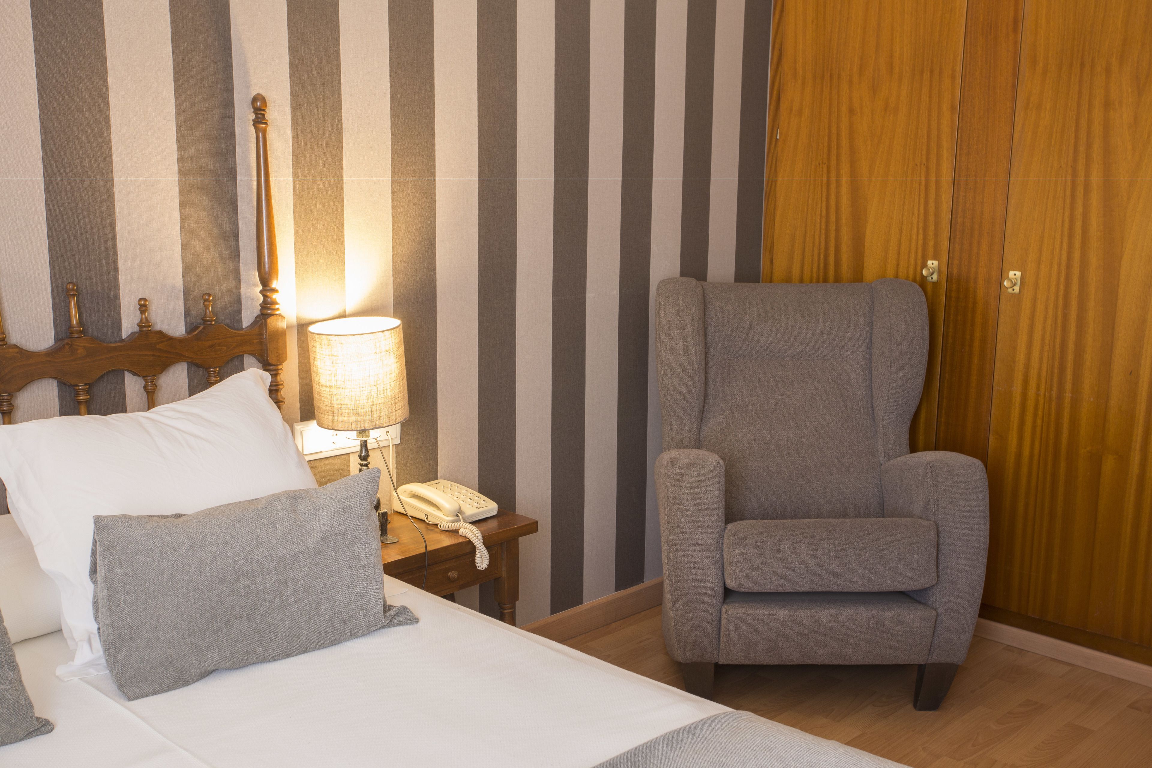 Habitació d'hotel amb un llit, una butaca gris, un armari de fusta i una taula de nit amb una làmpada i un telèfon.