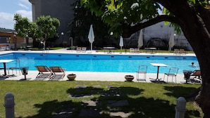 Una piscina rectangular con sillas de descanso y sombrillas, rodeada de césped y árboles.