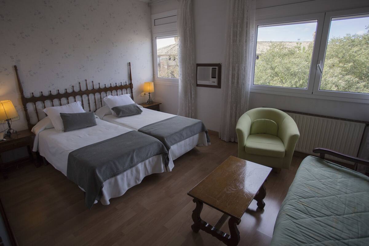 A bedroom with two beds, a chair, a table, and a window with a view.