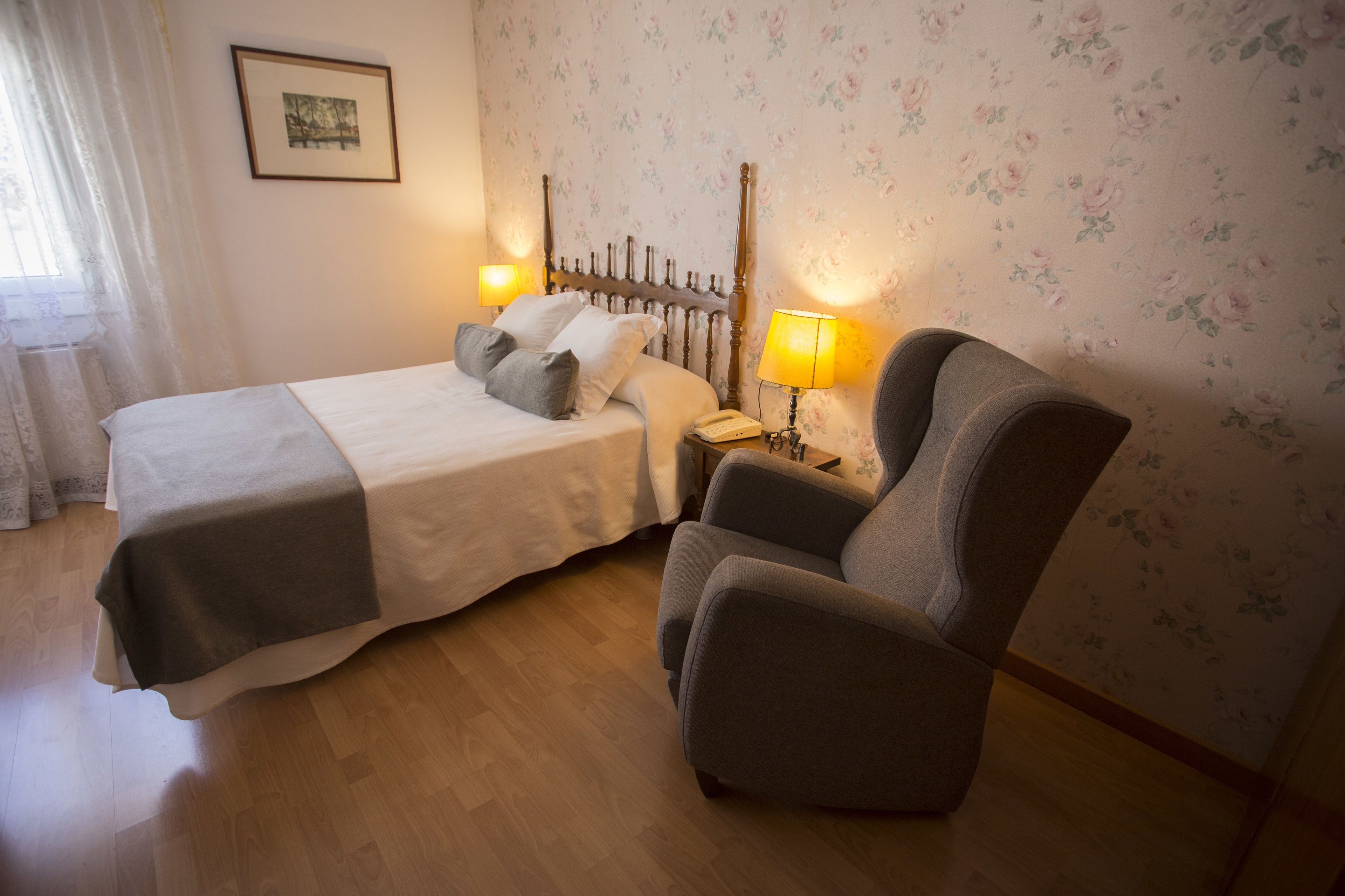 A bedroom with a bed, two bedside lamps, a chair, a nightstand, and a floral wallpaper.