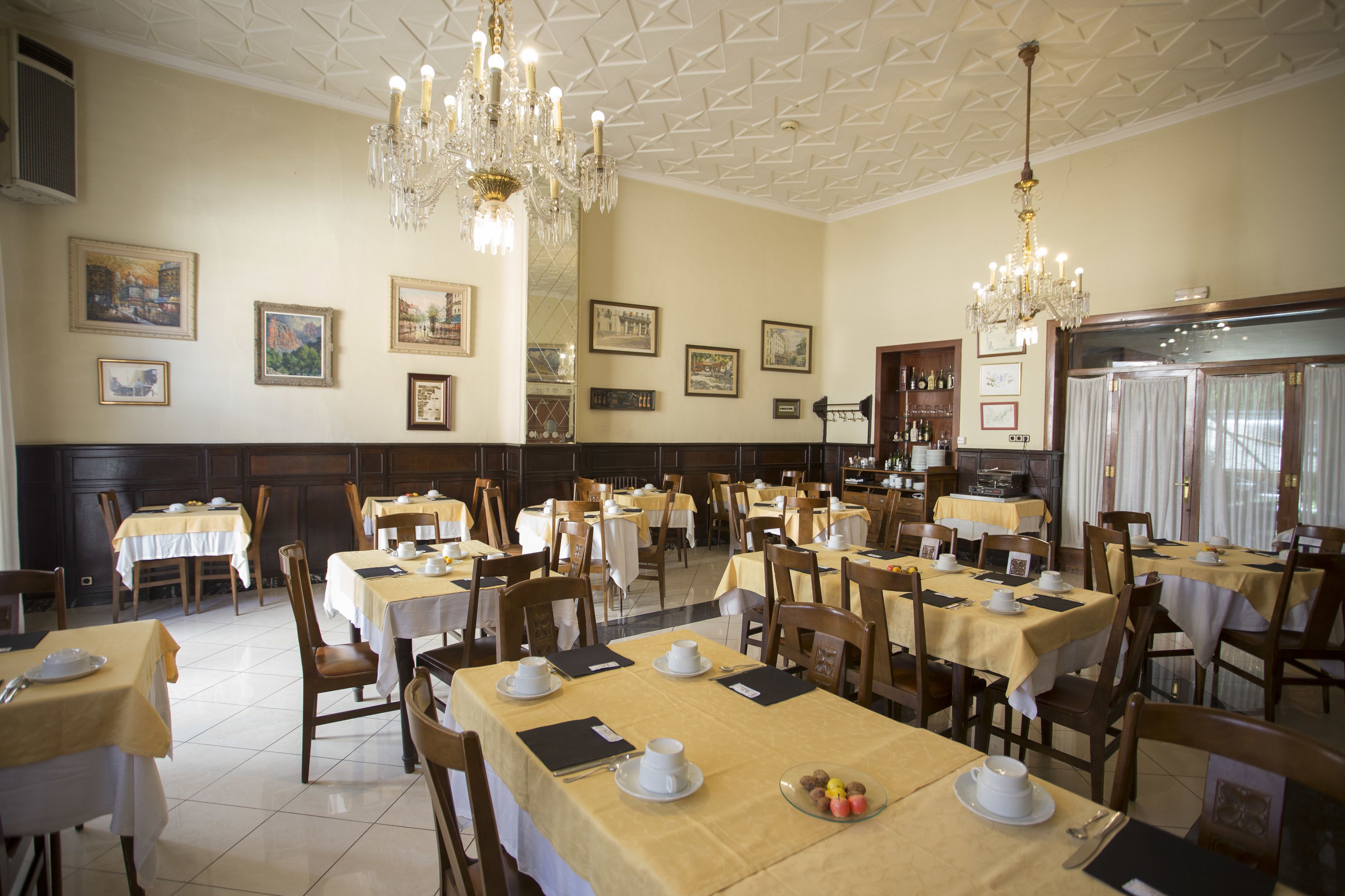 Un comedor amplio con varias mesas dispuestas para el servicio, elegantes candelabros y obras de arte enmarcadas en las paredes.