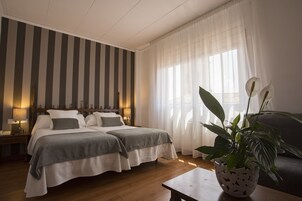 Habitación de hotel con dos camas, un ventanal con cortinas traslúcidas, suelo de madera y una planta en maceta sobre una mesa.