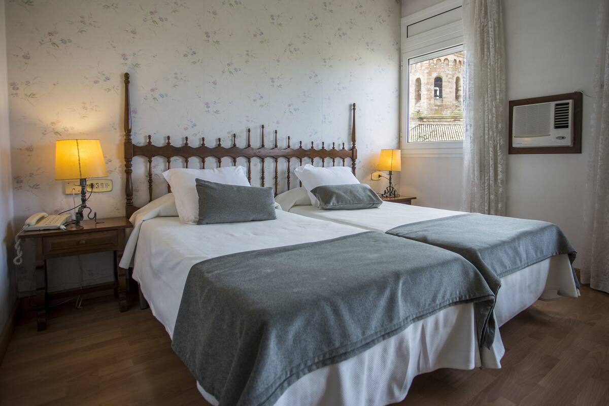 A double bed with two bedside tables, each with a lamp and a telephone. The room has floral wallpaper and a window with a view of a building.