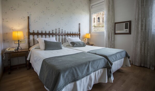 Una cama doble con dos mesitas de noche, cada una con una lámpara y un teléfono. La habitación tiene papel tapiz con motivos florales y una ventana con vistas a un edificio.