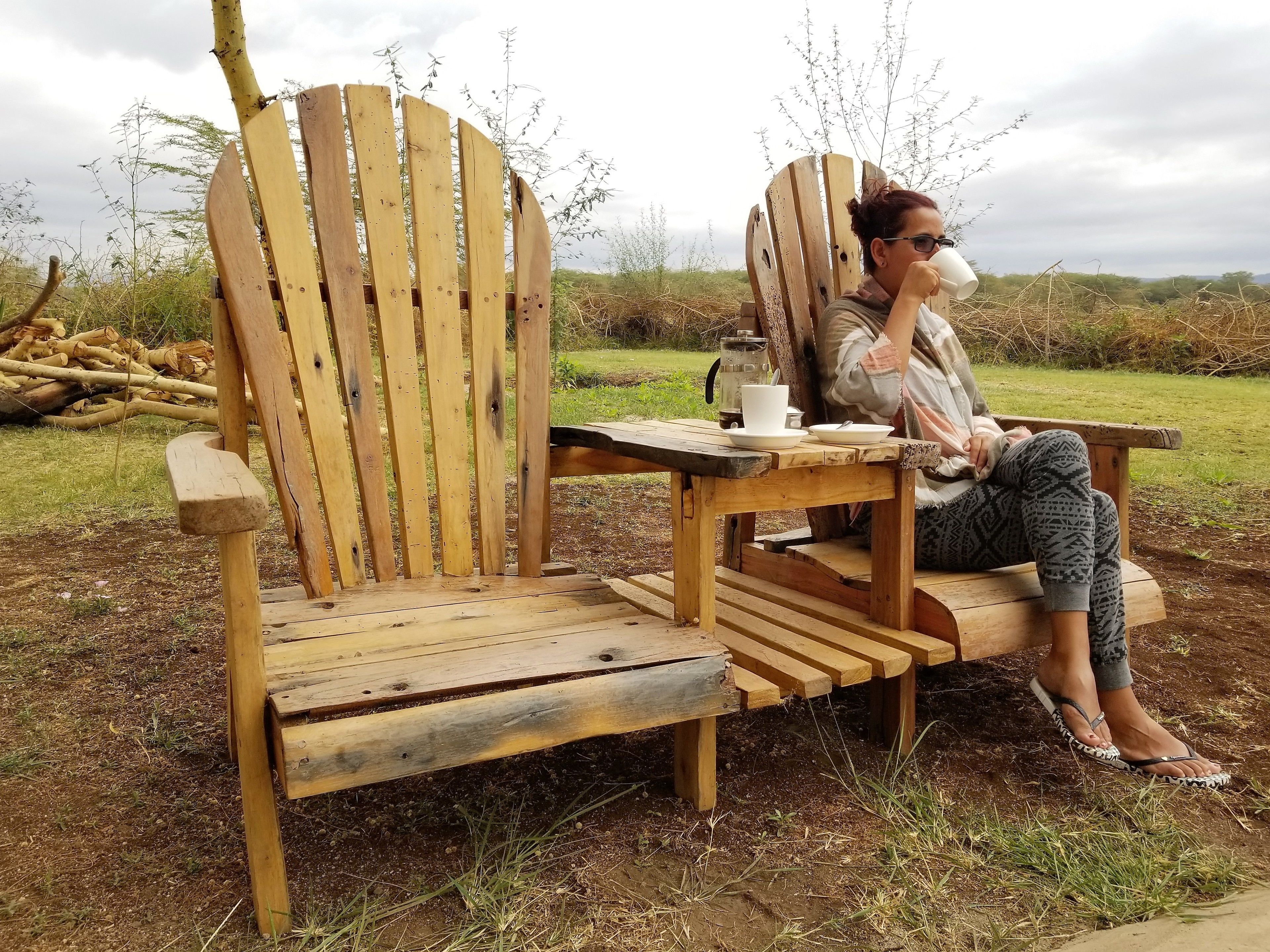 A wooden Adirondack chair with a table, a white cup, and a plate.