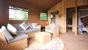 Ein Holzinnenraum mit einer Couch, einem Sessel und einem kleinen Holztisch. Ein Fenster bietet Blick auf Bäume, und eine Tür führt in einen Schlafbereich.