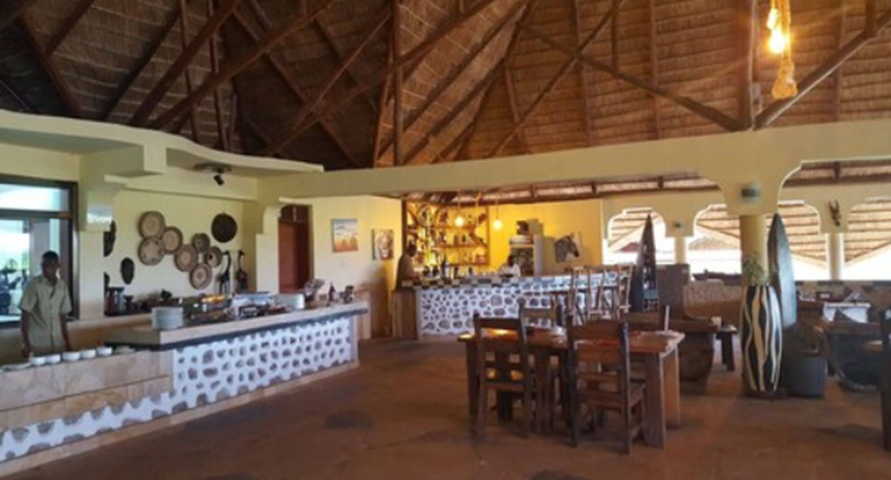 A spacious indoor area with a high ceiling and a thatched roof. There is a bar area with stools and a counter, and a dining area with wooden tables and chairs.