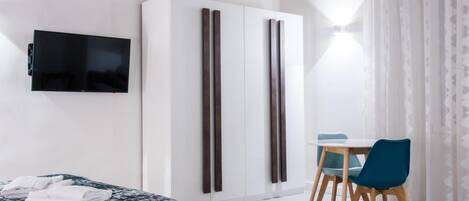 A modern hotel room with a bed, a flat-screen TV, a wardrobe, and a dining area with chairs.