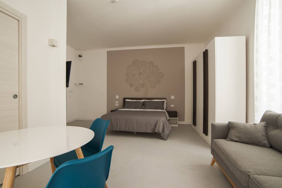 A modern bedroom with a bed, a gray sofa, a round table, and blue chairs.
