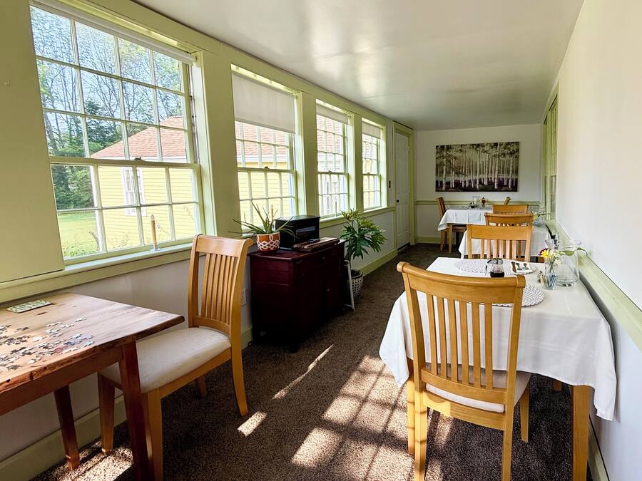 A dining area with wooden chairs and tables, large windows, and a painting on the wall.