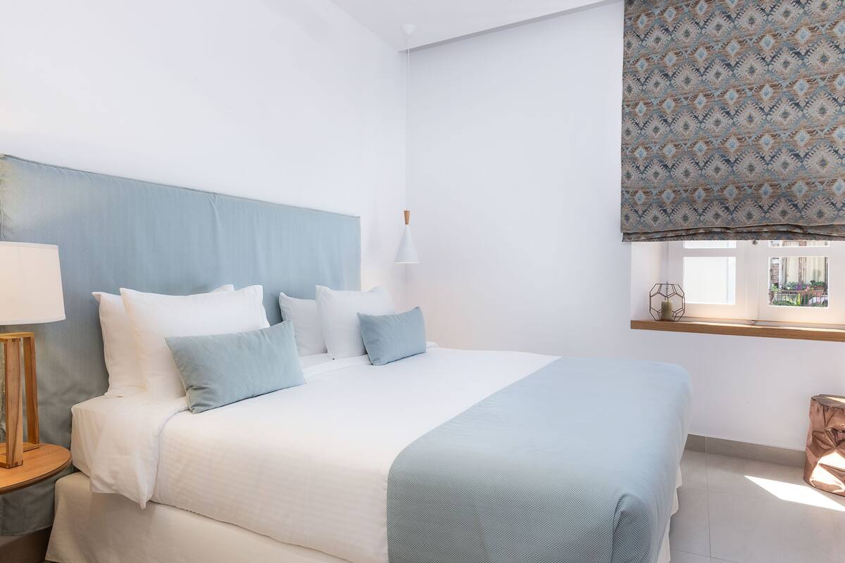 Un letto rifatto con lenzuola bianche e azzurrine, un comodino in legno con una lampada e una finestra con tende a motivi.