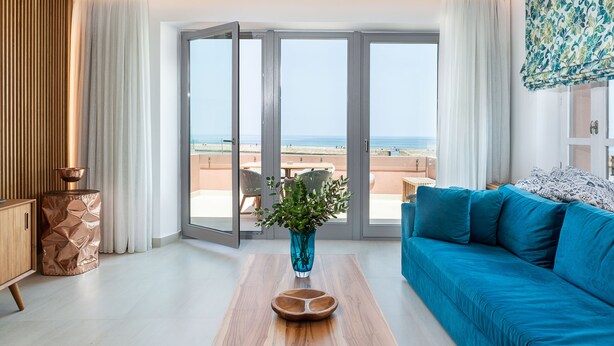 A modern living room with a blue sofa, a wooden coffee table, and a vase of flowers. There is a sliding glass door leading to a balcony with a view of the beach. - Avra Apartments Venetian Harbour (Chania)