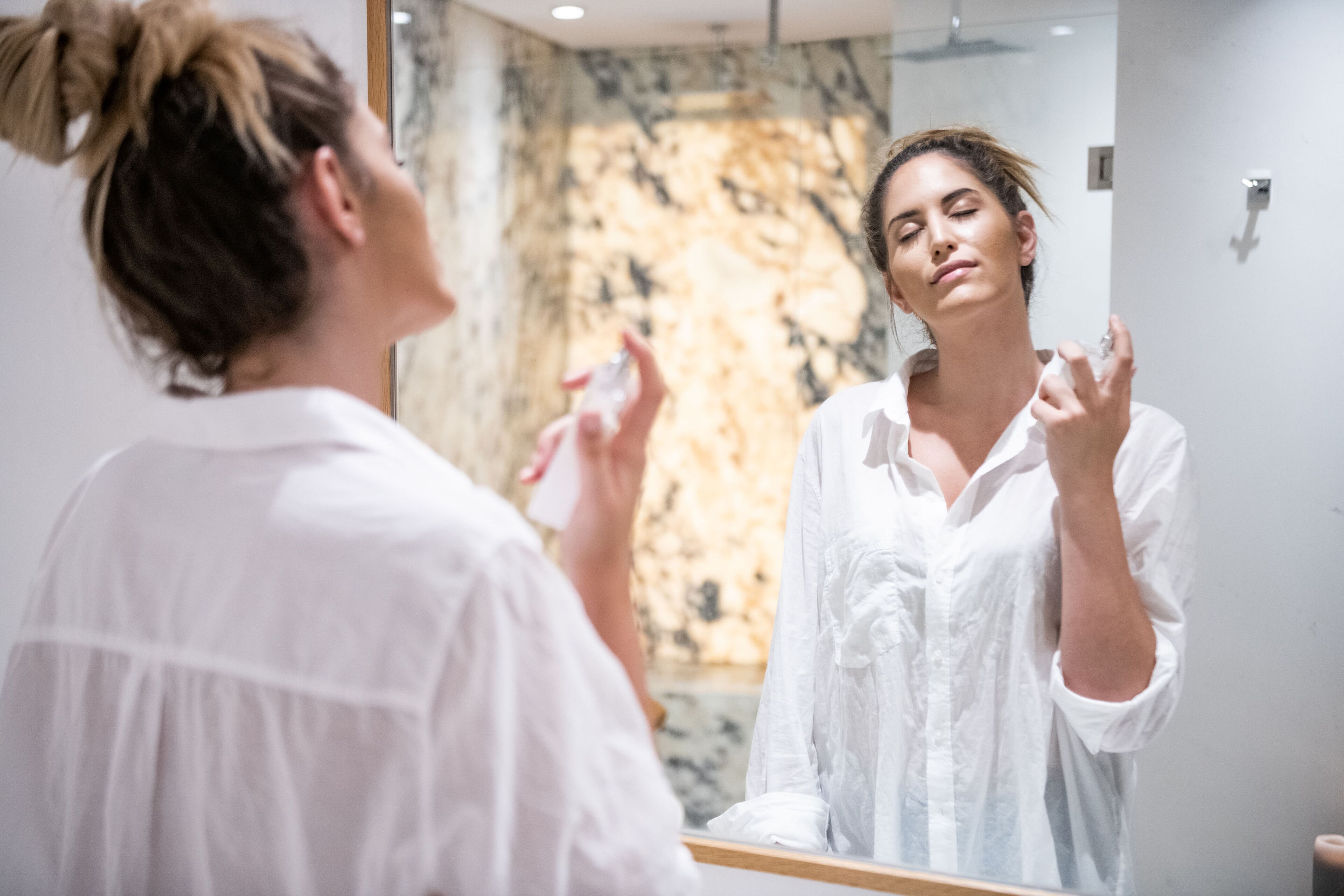 Una persona con camisa blanca de pie frente a un espejo en un baño con pared de mármol.