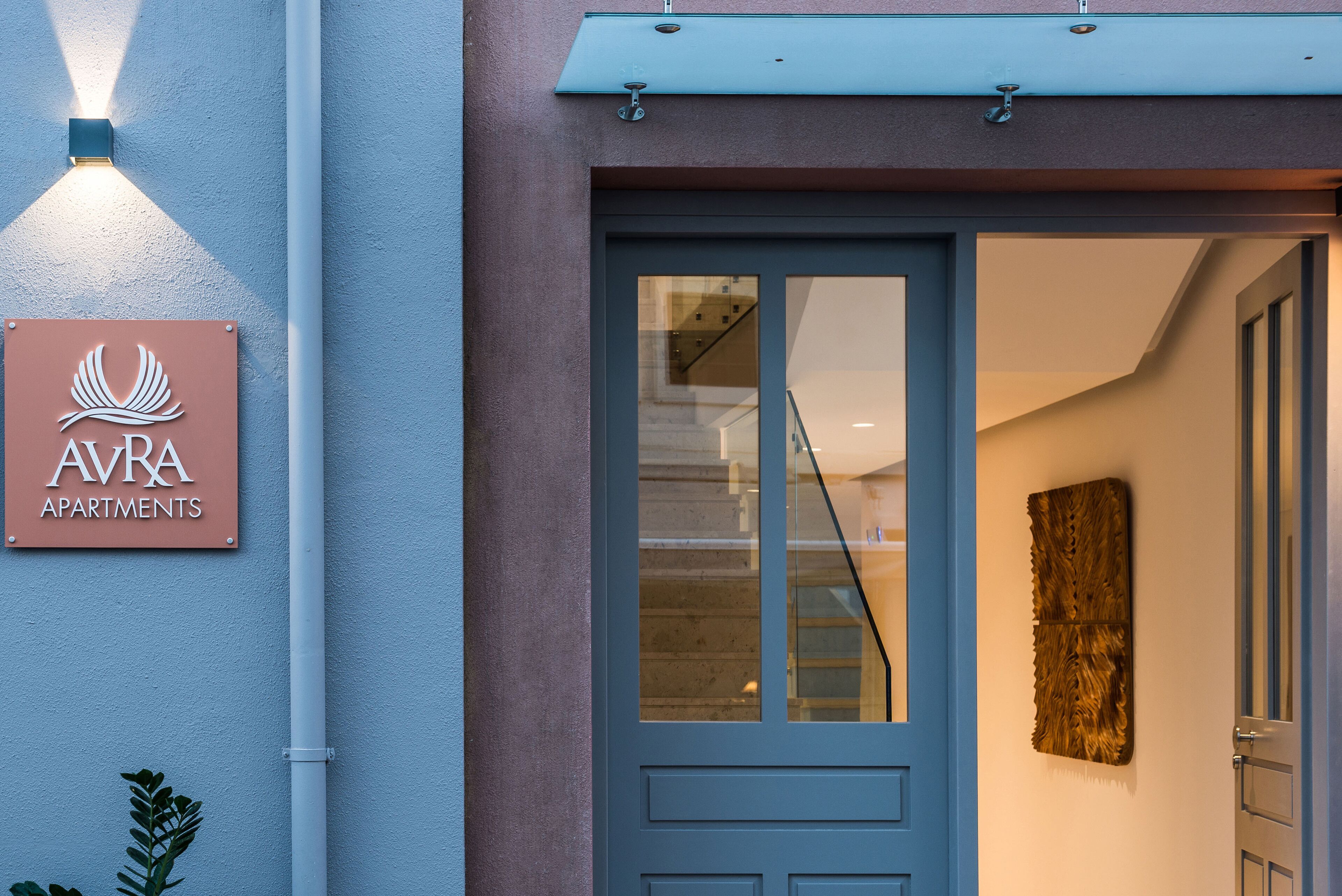 L’entrée d’un immeuble, avec une enseigne indiquant « APPARTEMENTS AVRA », une porte en verre et une œuvre d’art fixée au mur.