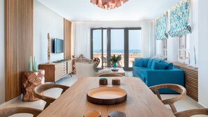 A modern living room with a wooden dining table, blue sofa, and large windows offering a view of the sea. - Avra Apartments Venetian Harbour (Chania)