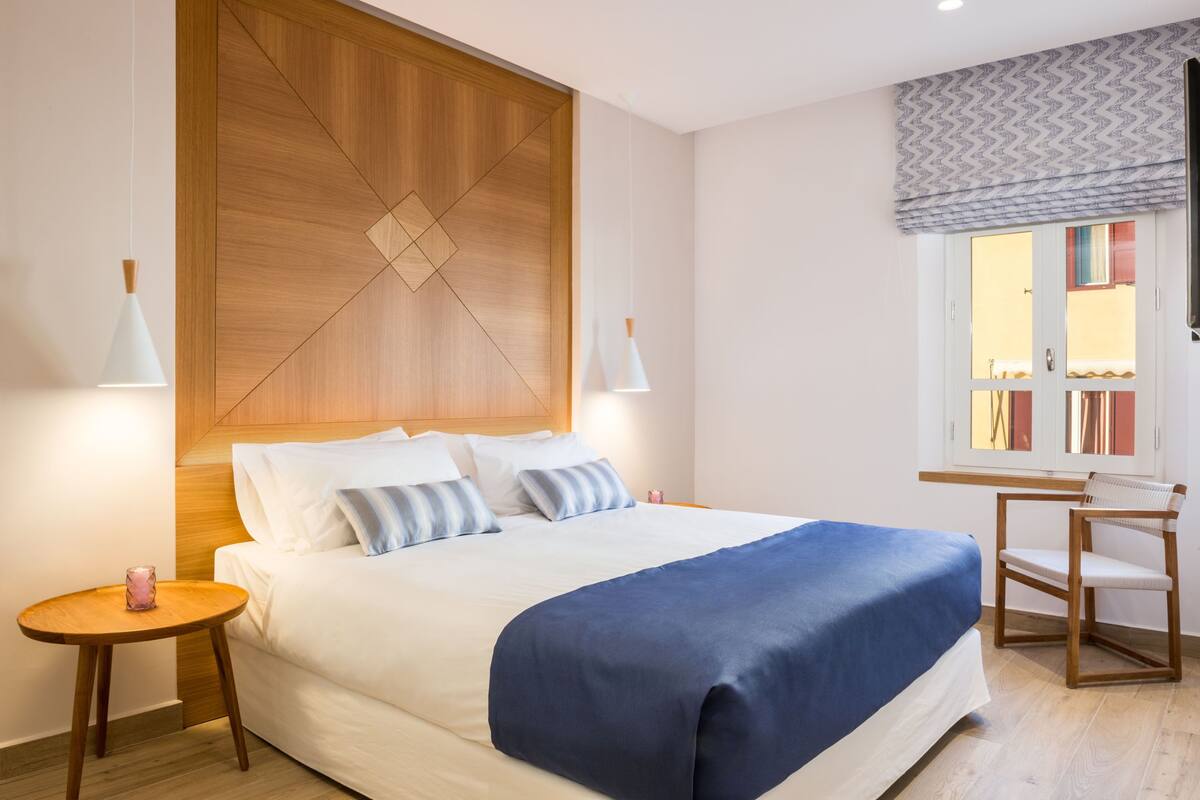 Un letto rifatto con cura, una coperta blu, due comodini con candele, una testiera in legno e una finestra con tende a motivi.