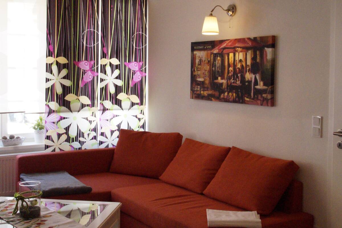 A living room with a red sofa, a coffee table, a painting on the wall, and a window with a curtain.