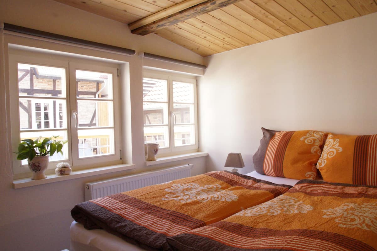 A bedroom with a bed, a window with a view of buildings, a radiator, and a plant on the windowsill.