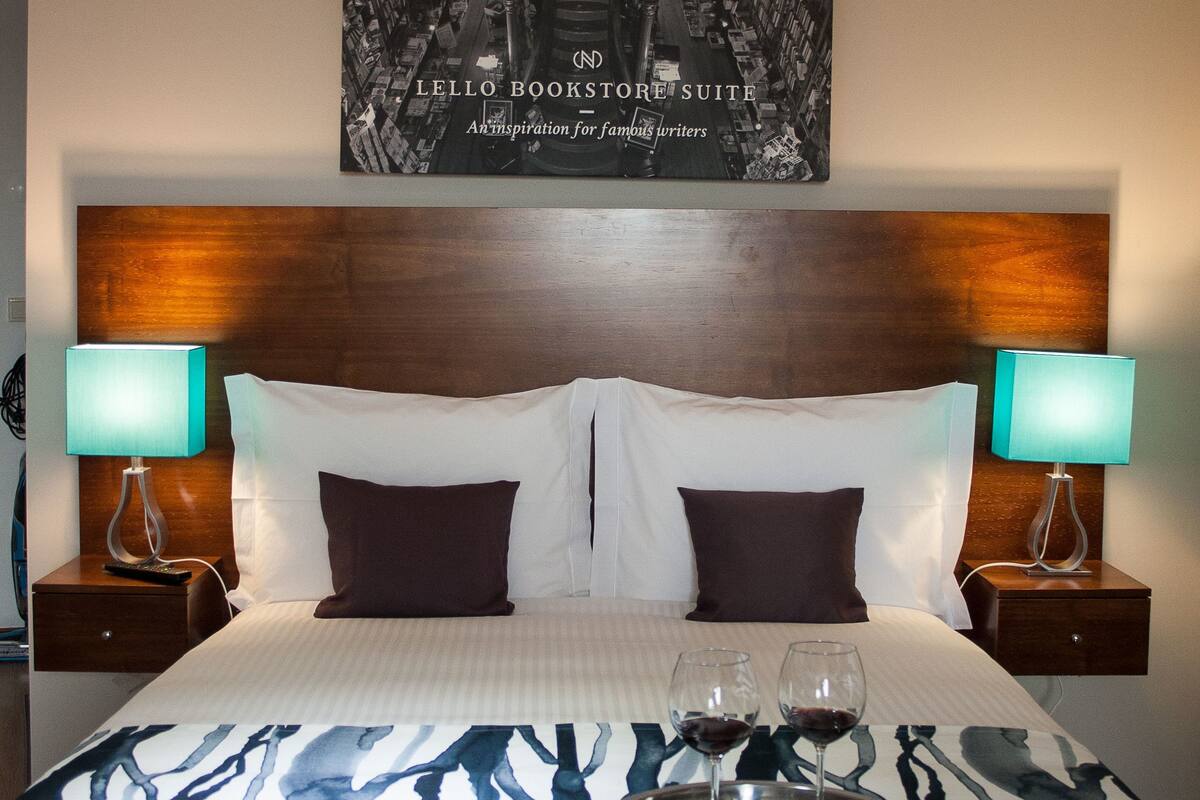 Habitación de hotel con una cama grande, dos mesitas de noche con lámparas, una bandeja con copas de vino y un cartel en la pared.
