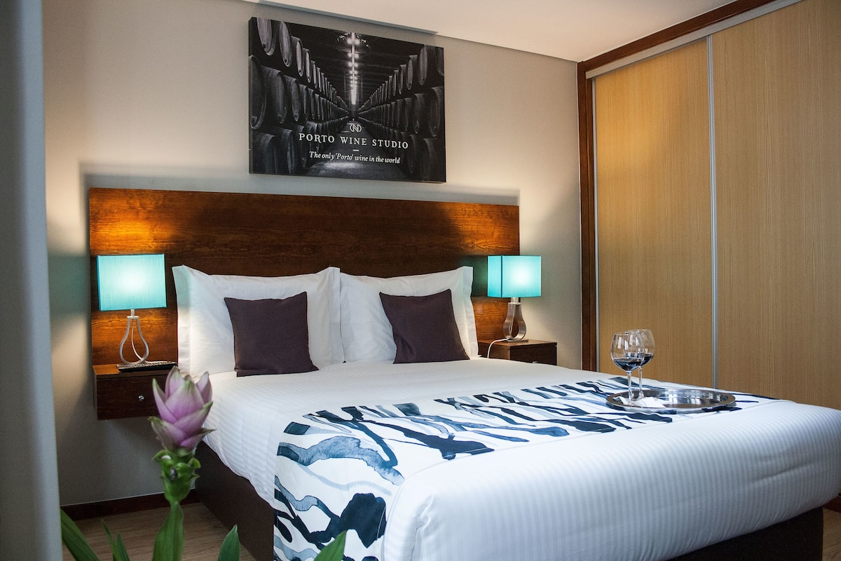 Habitación de hotel con una cama grande, dos lámparas de noche, una bandeja con una copa de vino y un cartel en la pared.