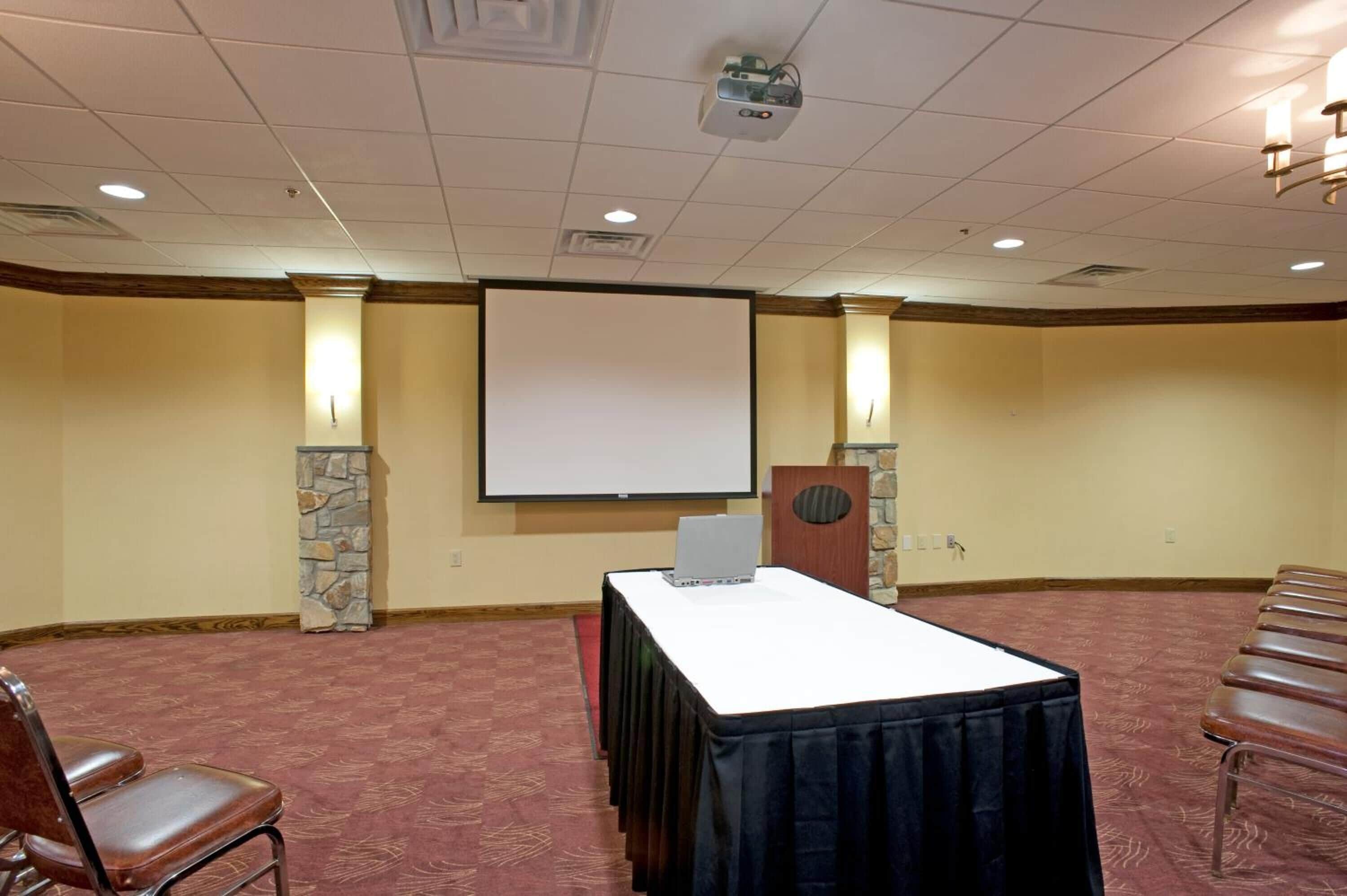 A conference room with a projector screen, a table with a laptop, stone pillars, and rows of chairs.
