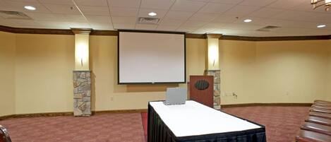 A conference room with a projector screen, a table with a laptop, stone pillars, and rows of chairs.