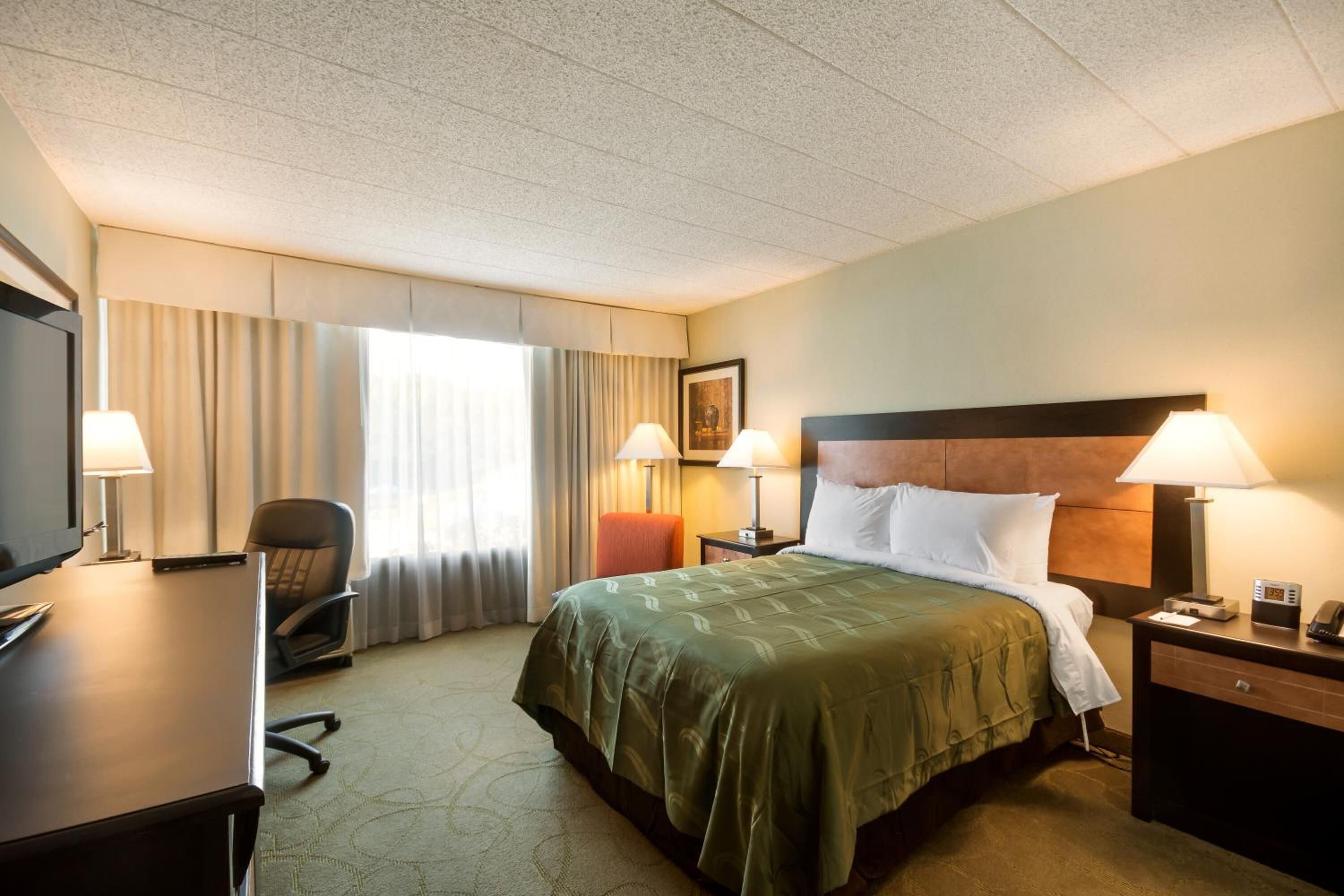 Habitación de hotel con una cama, dos lámparas de noche, un escritorio con una computadora, una silla y un ventanal con cortinas.