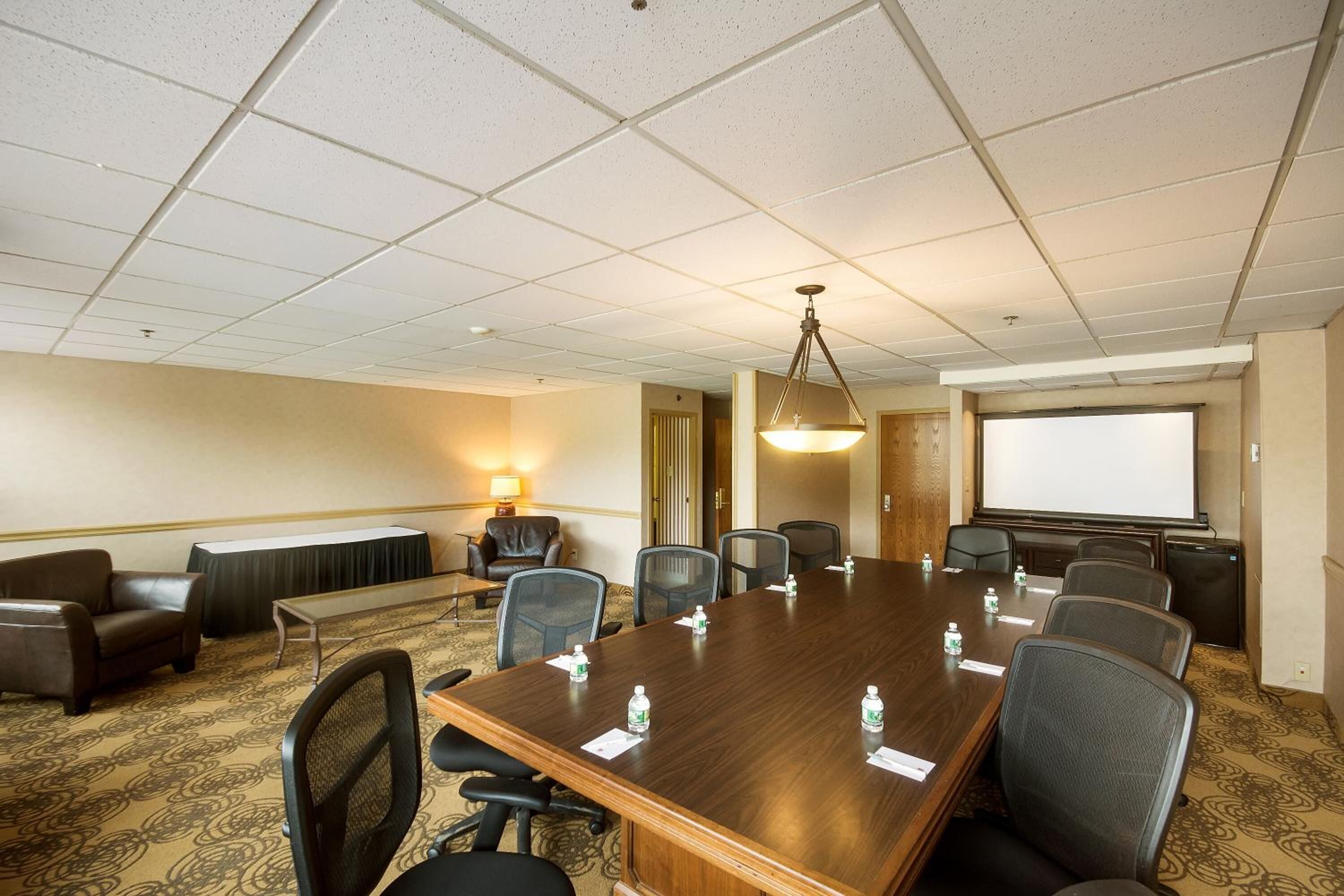 A conference room with a large wooden table, multiple chairs, a projector screen, and a carpeted floor.