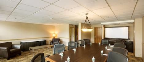 Una sala de conferencias con una gran mesa de madera, varias sillas, una pantalla de proyector y piso alfombrado.