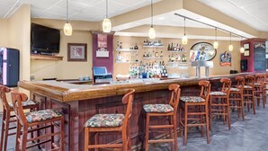 A bar area with wooden bar stools, a counter with a television above, and shelves stocked with bottles and glasses. - Chester Hotel & Conference Center (Exton)