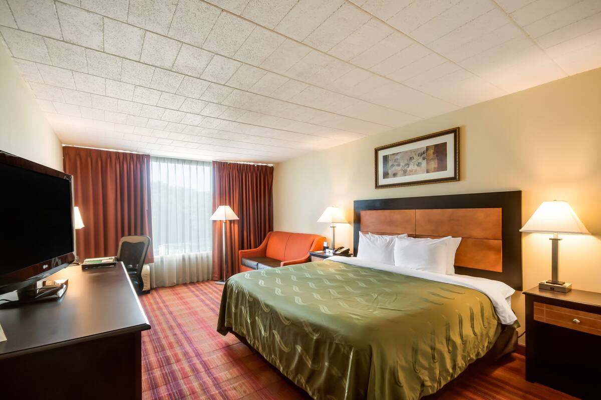 A hotel room with a bed, a desk with a television, a chair, a nightstand with a lamp, and a window with curtains.
