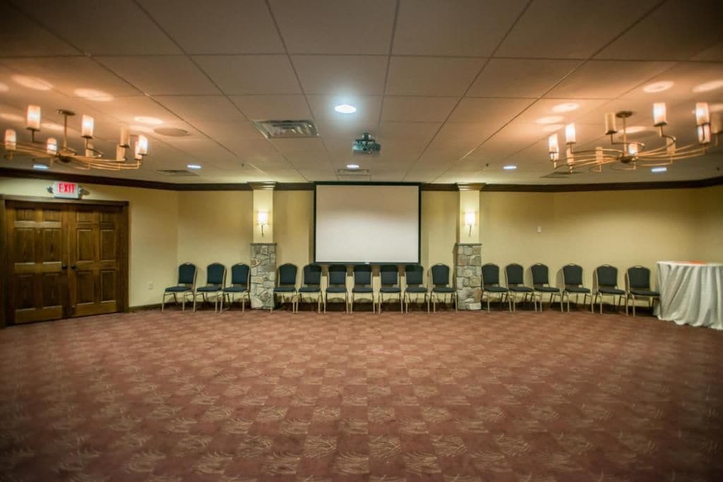A conference room with a carpeted floor, rows of chairs, a projector screen, and a wooden door.