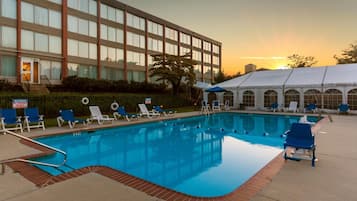 Una piscina con sillas de descanso y un área de estar techada, rodeada por un edificio con grandes ventanales.